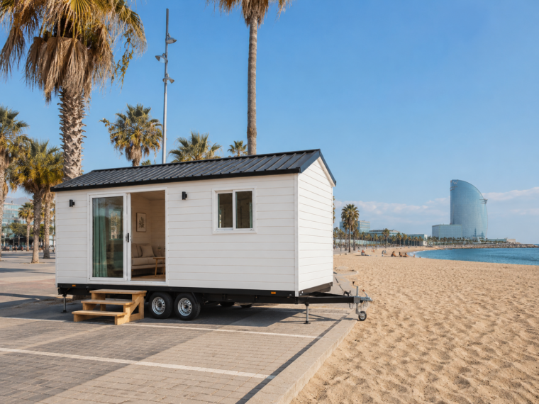 Biały domek mobilny na dwuosiowej przyczepie ustawiony przy piaszczystej plaży. Ma czarny dwuspadowy dach, przeszklone drzwi, małe okno i drewniane schodki. W tle widać palmy, morze i nowoczesny, wysoki budynek o zakrzywionej fasadzie.
