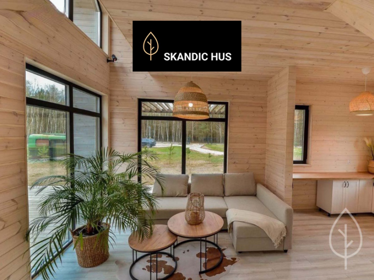 Jasne wnętrze domu Skandic 35 wykończone naturalnym drewnem, z wysokim sufitem i dużymi oknami w czarnych ramach. W salonie znajduje się beżowa narożna sofa, zestaw okrągłych stolików, pleciona lampa oraz duża palma w donicy. Po prawej widać niewielki blat roboczy z szafką, a przez przeszklenia otwiera się widok na zalesioną działkę.