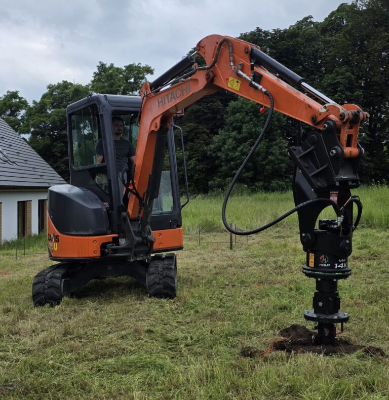 Minikoparka wyposażona w hydrauliczne urządzenie wkręca długi stalowy pal śrubowy w grunt na trawiastej działce. Na pierwszym planie widoczny jest wcześniej zamontowany pal zakończony kwadratową głowicą, przygotowaną do podparcia konstrukcji domu.
