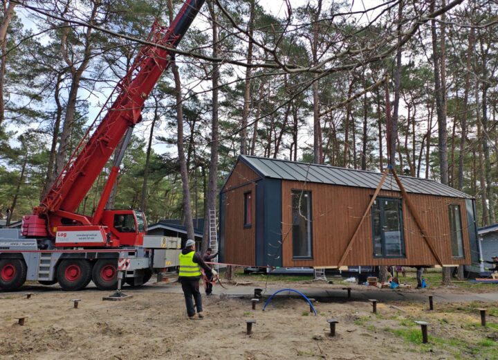 Drewniany domek modułowy unoszony przez czerwony dźwig podczas montażu na leśnej działce. Budynek ma pionową elewację z brązowego drewna, ciemne narożniki i dwuspadowy dach z blachy na rąbek. W ziemi widoczne są rozmieszczone pale wkręcane, przygotowane jako podparcie konstrukcji. Przy domku pracuje kilku montażystów.
